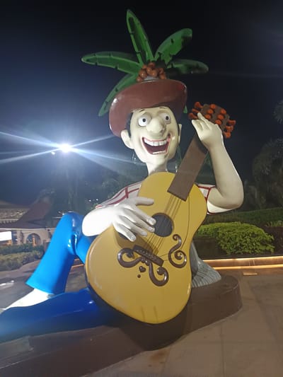 A large, colorful statue of a cartoon-like man playing a guitar is prominently featured at night in Kasarvarne, India. The statue depicts a fair-skinned man with exaggerated features, including a wide smile showing teeth, a large nose, and big eyes. He wears a brown hat adorned with green palm fronds and reddish-brown coconuts, a red and white horizontally striped shirt, and bright blue pants. His left hand is positioned on the neck of the yellow acoustic guitar, which has decorative brown swirl patterns and what appear to be brown frets or markers on the neck. His right hand rests on the body of the guitar as if strumming.

The statue is seated on a brown, low platform or base. In the background, the scene is dark, indicating nighttime. Bright artificial lights, possibly streetlights, cast a strong glare, illuminating parts of the statue and creating lens flares. Behind the statue, lush green bushes and trees are visible, along with parts of a building featuring arched structures, suggesting a public area, possibly a resort or park. The overall impression is cheerful and lively, consistent with the statue's grinning expression and musical theme. No visible text is present in the image.