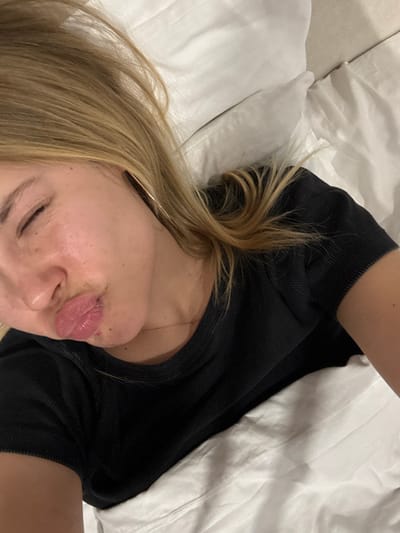 A woman with blonde hair is lying in bed, partially under white sheets. She's wearing a black top and has her eyes closed. Her lips are puckered slightly. The photo is taken from an above, angled perspective, emphasizing her face and upper body against the white bedding.