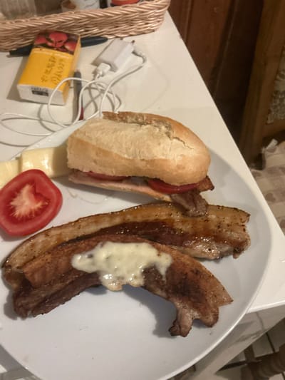 A plate of food is on a white table. The plate contains a sandwich, three pieces of pork belly, a slice of tomato, and a piece of cheese. The pork belly is topped with melted cheese. In the background, a wicker basket and a phone charger are visible. The image appears to have been taken in a domestic setting, possibly a kitchen or dining room, in Wenzenbach, Germany.