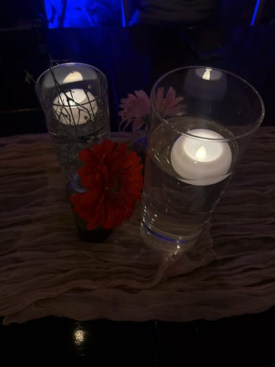 The image presents a dimly lit, interior scene featuring two cylindrical glass vases positioned horizontally. One vase contains a white, battery-operated candle and a pink flower. The other vase contains a similar white candle and a vibrant orange gerbera daisy. Both vases appear to have water within them. A beige, textured fabric drapes behind the vases. To the left, blue lighting illuminates what appears to be a black, tiled surface. The overall setting evokes a decorative ambiance, possibly for an event or gathering. While the image's origin is stated as Santiago de Querétaro, Mexico, there is no explicit indication within the image itself to confirm this location.