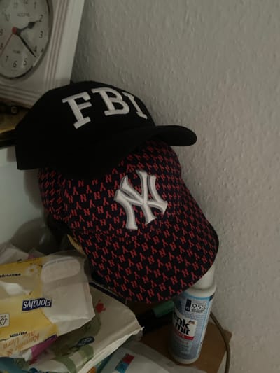 A close-up shot captures two baseball caps stacked on a shelf. The top cap, black with white lettering, displays the letters "FBI". Beneath it, a second cap features a red and black pattern of "NY" logos, with a white "NY" emblem on the front. To the left of the caps, a white analog clock with black numbers and hands is visible. In the foreground, to the left of the caps, a package of baby wipes with yellow and white packaging is partially visible. To the right of the caps, a blue and white bottle of cleaning product is present. The background consists of a textured, off-white wall. The lighting appears to be artificial and subdued, casting shadows on the wall. The scene is indoors, likely in a domestic setting. There is no information to place this scene in the city of Kassel, Germany.