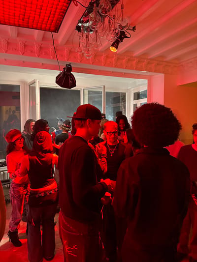 The image shows a group of people standing in a room with red lighting. The location is a building in Palaiseau, France. There are windows in the background with a white window frame. There is a hanging light and a softbox lighting rig above the group of people.
