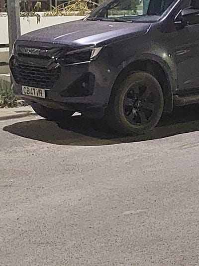 A dark-colored SUV is parked on a paved street in São Teotónio, Portugal. The vehicle's front grille and headlights are visible, along with a license plate that reads "CB47VR". The car's tire and fender are also in focus. The background shows a wall with some vegetation and what appears to be a utility pole. The street surface is textured and appears to be asphalt. No people are present in the image.