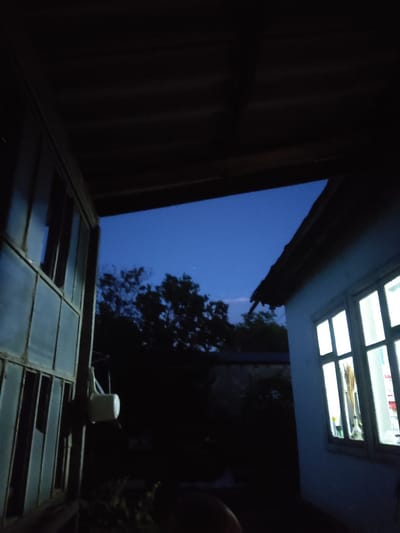 This media file depicts an outdoor scene at dusk or early night. The view is looking upwards and outwards from an enclosed or partially enclosed space, possibly a verandah or a covered patio with a corrugated roof visible on the left. Dominating the upper part of the frame is a window with multiple panes, illuminated from within, suggesting it is inside a building. The light from this window casts a glow on the ceiling structure around it.
Below this upper window, a large opening reveals a deep blue twilight sky, with a few trees silhouetted against it. The sky appears clear. In the foreground, another window structure with multiple panes is visible, partially obscured by darkness. Some of these panes appear to be open or missing, allowing a view into the darker space beyond.
On the ledge of this foreground window structure, a white plastic bucket with a handle is placed. The overall impression is one of a somewhat rustic or utilitarian setting, perhaps a backyard or a common area. The time of day is clearly evening, as indicated by the dark sky and the artificial light from the upper window. The location cues, such as the corrugated roof and the style of the window frames, could be typical of rural or less developed areas. No people are visible in the frame. The emotion conveyed is one of quiet stillness, perhaps a sense of transition between day and night. There is no visible text.