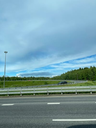 The image depicts a view from a moving vehicle on a highway in Medik, Russia. The sky is blue with streaky clouds. A line of trees borders the highway, separated by a green grassy area and a metal guardrail. The highway has multiple lanes separated by white lane markings. There is one dark-colored vehicle visible on the highway, at the far left, behind the guardrail. A tall light pole is visible in the lower left corner of the image.