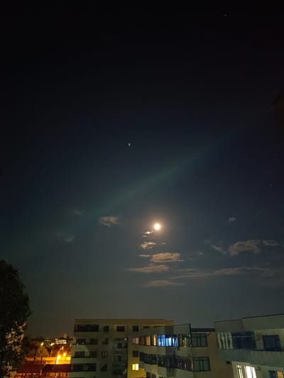 The image captures a nighttime view of the sky and city buildings in Târgu Jiu, Romania. The sky is dark blue, with the moon prominently visible near the center of the frame, appearing as a bright, luminous orb. Several small clouds are scattered across the sky, some illuminated by the moonlight. A faint, luminous band stretches across the sky from the moon towards the upper right quadrant. Tiny points of light, likely stars or distant celestial bodies, are visible in the upper portion of the sky.

Below the sky, the tops of several city buildings are visible. The buildings are multi-story structures with visible windows, some of which are illuminated with artificial light. A large tree with dark foliage is present on the left side of the frame, partially obscuring the view of the buildings and streetlights below. Streetlights cast an orange glow on the scene, particularly in the lower left portion of the image. The overall impression is a quiet, nocturnal urban landscape under a moonlit sky.