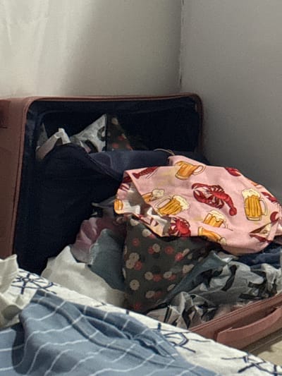 An opened, pink suitcase is filled with various clothing items. The clothing includes a pink garment with a repeating pattern of lobsters and beer mugs, a dark blue garment, a gray and white patterned fabric, and other folded and crumpled textiles. The suitcase is positioned on a surface, possibly a floor or bed, and the background consists of a plain, light-colored wall. There are no people visible in the image. The scene is indoors, and the lighting appears to be artificial. The image does not provide any information about the location in the city of Asprovalta, Greece.