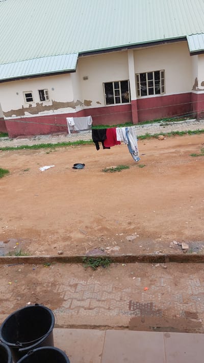 The image depicts an outdoor scene in New Nyanya, Nigeria, featuring a building with a light green corrugated roof and cream-colored walls with a red trim. The building has several windows, some with bars and others open. In front of the building, clothes are hanging on a clothesline, including a grey shirt, black pants, a red shirt, and light blue pants. There are also some clothes lying on the ground, one white and one dark colored. The ground is mostly dirt with some patches of grass and scattered debris. In the foreground, part of a paved area is visible with two black buckets. The overall impression is one of a residential or institutional setting with everyday activities taking place outdoors.