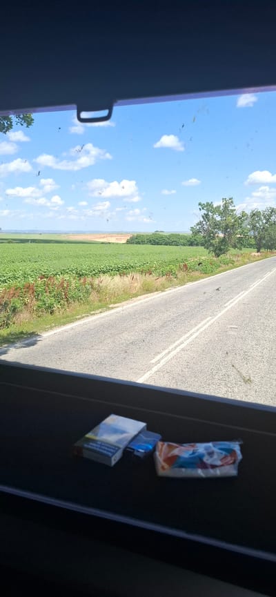 This is a photograph taken from inside a vehicle, looking out the window. The scene depicts a rural landscape with a paved road running through it. The road is gray with white lines indicating lanes. To the left of the road is a field of green crops, possibly sunflowers, stretching towards a distant line of trees. Above the field is a bright blue sky with scattered white clouds.
On the right side of the image, inside the vehicle, there are two items placed on a dark surface. The top item is a small, crinkled plastic bag containing an orange and blue object. Below this is a pack of cigarettes. The cigarette pack is mostly white with blue and orange accents, and some text is visible on it, though it is not legible.
The time of day appears to be daytime, with bright sunlight casting shadows and illuminating the scene. The weather seems to be clear and sunny. The overall impression is one of a journey through the countryside on a pleasant day. The presence of the cigarette pack suggests a passenger or driver might be smoking. The image is taken from an elevated perspective, perhaps from a car or bus window. The exact location is given as Zgalevo, Bulgaria.