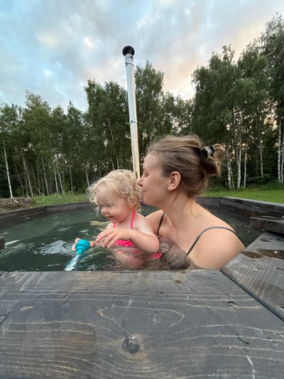 The image shows a mother and her young child in a wooden hot tub. The mother, with her hair in a bun, is leaning down and kissing the child's forehead. The child, with blonde curly hair, is wearing a pink swimsuit and holding a blue toy. They are both in the water, which appears to be murky green. The hot tub is made of dark, weathered wood. In the background, there is a lush green forest with birch trees. The sky is cloudy and overcast, suggesting either early morning or late evening. The overall mood of the image is warm and intimate, conveying a sense of affection between mother and child. The location context provided is Ryzhkovo, Russia. There is no visible text in the image.