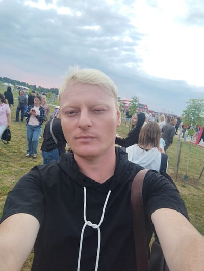 The image is a selfie taken by a light-skinned man with short, bleached blonde hair, wearing a black hooded sweatshirt with white drawstrings. He has freckles on his arms. He is standing outdoors in an open grassy area with several other people in the background. The sky is overcast with dramatic cloud formations. In the distance, there are tents and structures that suggest an outdoor event or festival. Trees are planted in rows, some with visible trunks and minimal foliage, indicating they are young. The ground is covered in green grass. The overall atmosphere suggests a public gathering on a cloudy day. The location is identified as Buinitski Sel'ski Sovet, Belarus, which is likely an outdoor park or event space within that administrative region.