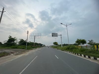 The image depicts a road in Purbachal, Bangladesh under a cloudy sky. The road is asphalt, marked with white lane lines and flanked by black and white curbs. On the left side of the road, there is a puddle of water near overgrown vegetation and utility poles. A billboard frame stands to the left, and a road sign indicating "Underpass" and "Kanchan Bridge" is visible in the middle of the road. Streetlights line the road on both sides. In the distance, a single motorcyclist is visible. The environment is characterized by green vegetation and an overcast sky.