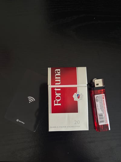 An overhead, close-up shot shows a black table with a pack of Fortuna cigarettes and a red Blink lighter. The pack of Fortuna cigarettes is vertical and to the left of the lighter. The pack is mostly white with a red stripe that has "Fortuna" written in white lettering. To the left of the cigarettes, there is a black access card with a wifi symbol on it, and the word "openkey" in white lettering near the bottom. The red lighter is to the right of the cigarettes, and is lying on its side. The lighter has "Blink" written on it, and a warning label that says "WARNING: KEEP AWAY FROM CHILDREN." The image appears to be taken indoors. The location in San Francisco, United States of America is not visible in the image.