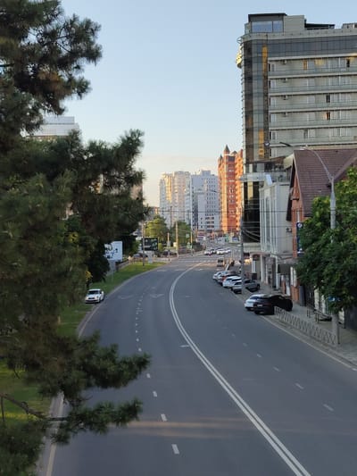 The image shows a wide, empty asphalt road curving through a city under a clear blue sky. On the left, a large evergreen tree with dark green needles partially obscures the view. The road has two white dashed lines dividing it into lanes, and double solid white lines on the right side. On the left side of the road, there is a white car parked on the grass verge, with a billboard in the background. On the right side, a row of parked cars lines the pavement, and a modern, multi-story building with a glass facade is visible behind them. Further down the street, several other apartment buildings of varying heights and architectural styles are visible, bathed in the warm light of what appears to be late afternoon or early morning sun. The buildings exhibit a mix of modern and older designs, with some featuring balconies and visible air conditioning units. A brown-tiled roof building with a pitched roof is situated closer to the foreground on the right. The scene appears tranquil, with minimal human activity evident. The overall impression is a contemporary urban environment with a touch of nature provided by the trees. While the exact location within Novomoskovsk, Russia, cannot be determined from the visual cues alone, the presence of Russian-language billboards or signs would have provided a stronger geographical indicator. However, the architectural style of the buildings, particularly the multi-story apartment blocks, is consistent with urban development found in many Russian cities.