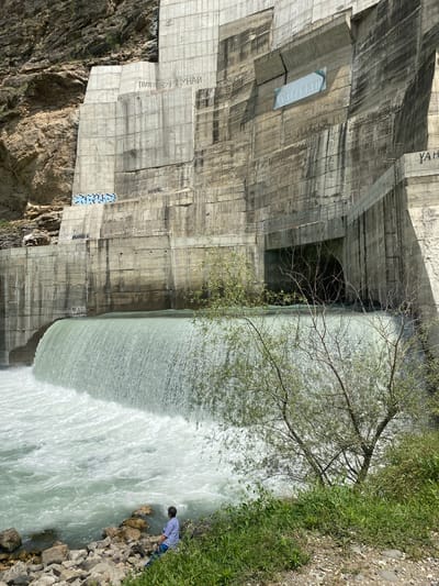 An outdoor, vertical, eye-level shot captures a man sitting on a rocky bank beside a wide, powerful waterfall cascading over a massive, concrete dam. The dam dominates the background, its textured surface marked with weathered patches, streaks, and graffiti. Cyrillic inscriptions and symbols are visible on the dam's face. A large, dark opening, likely an outflow tunnel, is situated in the center of the dam. The water is a milky, turquoise color, churning and frothing as it falls and flows into the river below. Greenery, including bushes and trees, grows along the bank in the foreground and on the dam's slopes. The man, viewed from behind, wears a blue shirt and dark pants, facing the waterfall. The surrounding environment appears to be a mountainous, natural setting, with rocky cliffs visible behind the dam. The location is identified as the Chirkey Dam in Untsukul', Russia.