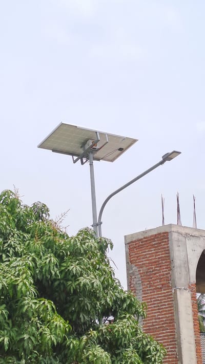 A solar-powered street light stands tall against a pale sky, its panel angled upwards to capture sunlight. A curved metal arm extends from the main pole, supporting a modern LED street lamp. To the left, a lush green tree with dense foliage partially obscures the view. To the right, the corner of a brick building under construction is visible, with concrete framing and exposed rebar. The scene appears to be an outdoor environment, possibly in a semi-urban or developing area.
