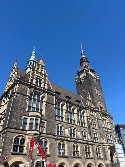The image shows the Elberfeld Town Hall in Wuppertal, Germany, from a low-angle perspective. The building's facade is made of dark grey stone with numerous windows. Two towers are visible, one with a green spire and the other with a darker spire. A red mobile crane is positioned near the building's base. The sky is clear and blue.