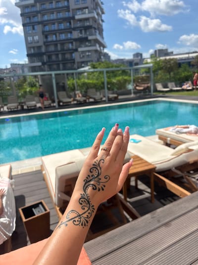A woman's hand with a henna tattoo and manicured fingernails is held up in the foreground, obscuring a portion of the scene. In the background, a swimming pool is visible, surrounded by lounge chairs and lush greenery. A modern, multi-story building stands prominently behind the pool. The sky is blue with scattered clouds. The location is the Kâğıthane district of Istanbul, Turkey.