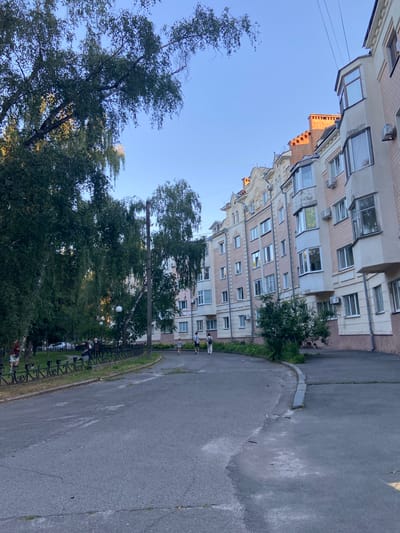 This media file captures an exterior view of a residential building in Nyzhni Mlyny, Ukraine. The building, with its light-colored facade and numerous windows, appears to be several stories high. It is a daytime scene, with clear blue skies suggesting fair weather. The building is situated next to an asphalt road, with a patch of green grass and trees bordering the road and the building. A dark metal fence separates the grass from the road in some areas. In the foreground, several people are visible, some walking along the road or sidewalk, while others are near the fence, possibly sitting or standing. One person is seen on the road wearing a light-colored shirt and shorts. Another person, also in light clothing, walks further down the road. In the middle ground, closer to the building and behind the fence, there are more individuals, some appearing to be in casual attire. The presence of trees and grass indicates an urban or suburban setting with some greenery. The lighting suggests it is likely late afternoon or early evening, as the sun casts a warm glow on the upper portion of the building, highlighting some architectural details. No visible text is present in the image. The overall impression is a peaceful, everyday scene in a residential neighborhood.