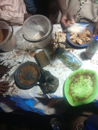 In an outdoor setting, likely in Tundun Wada West, Nigeria, a group of individuals are engaged in preparing food, possibly for a communal meal or a street vending operation. The scene is cluttered with various food preparation items. Two translucent plastic buckets are visible, one containing what appears to be finely ground grain or flour, and the other empty and slightly grimy. A blue bowl holds pieces of cooked food, possibly yam or cassava, which are being seasoned. A person's hands are adding a white powdery substance, likely spices or seasonings, to the bowl.

Other objects include a plastic water bottle, partially filled with a yellowish liquid, and a black plastic bag containing more of the cooked food or ingredients. A dark-colored plate with a reddish-brown residue, likely spices, sits beside a sponge-like object and a piece of brown, dried material. A bright green plastic plate, also with residue, is positioned towards the foreground.

The ground beneath the makeshift table appears to be dirt, with some blue plastic sheeting partially covering the area, suggesting an informal or temporary setup. The lighting suggests it is daytime, though the exact time is difficult to determine. The overall impression is one of active food preparation in a rural or semi-urban environment. No discernible text is present in the image.