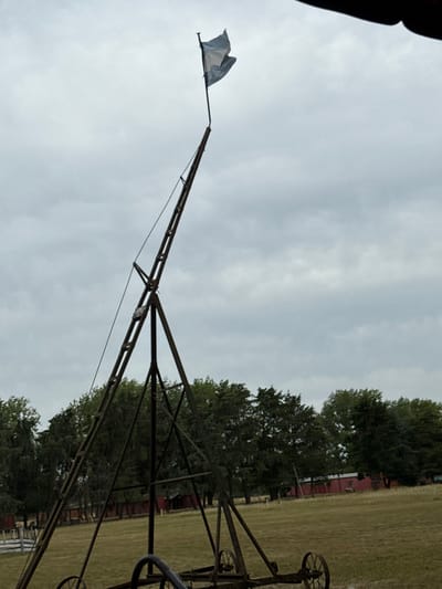 A tall, vintage metal structure with a flag at its peak stands against a cloudy sky. The structure features a lattice framework and appears to be on wheels, suggesting it could be moved. In the background, there is a field and some trees, partially obscuring a red fence. The overall scene has a rural or historical feel.