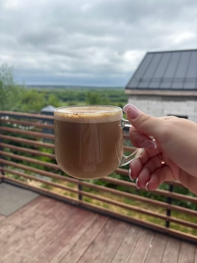 A clear glass mug filled with coffee is held by a woman's hand. The mug has a handle and is positioned in the center of the frame. The coffee has a frothy, light brown crema on top. The woman's hand is visible on the right side of the mug, with manicured fingernails painted with a white French tip and small pink speckles. A gold ring is on her ring finger. The background suggests an outdoor setting. A wooden deck or balcony is in the foreground, with horizontal wooden railings. Beyond the railings, there is lush green foliage and trees. In the upper right portion of the background, a portion of a building with a dark, slanted roof and light-colored brick walls is visible. The sky above is overcast with gray clouds. The overall scene implies a relaxed moment, possibly on a balcony or porch overlooking a natural landscape. The location is within the city of Semiluki, Russia.