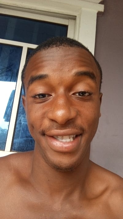 A head-and-shoulders portrait captures a young man with a dark complexion, smiling broadly at the camera. He has short, dark hair, a slight mustache, and stubble on his chin. His lips are shiny, and he shows his upper teeth in a wide, cheerful grin, with his eyes slightly squinted from the smile. The man is shirtless, revealing his bare shoulders and upper chest. A distinct lighter patch of skin is visible on his left collarbone area. The background features a window with white frames and blue-tinted glass on the left side, possibly reflecting an outdoor scene or sky. On the right, there is a plain, light-colored wall or structure. The lighting appears bright and natural, indicating daytime. The image conveys a warm and friendly emotion, set in Agburu Uke, Nigeria. There is no visible text.