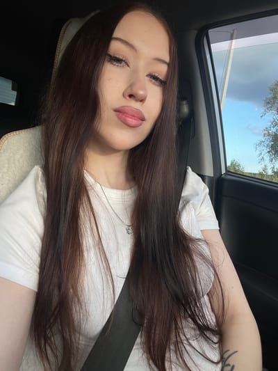 This is a close-up, rotated portrait of a young woman inside a car. She has long, dark brown hair that is spread across her chest and shoulder. Her face is turned towards the camera, with her eyes looking slightly upward. She has noticeable makeup on her eyes, with defined eyelashes and eyeliner, and her lips are painted a soft pink. A delicate silver necklace with a small charm is visible against her white t-shirt. The background shows the interior of a car, with a seatbelt visible. Through the car window and sunroof, a cloudy sky with some trees can be seen, suggesting it is daytime. The overall mood appears relaxed and casual.