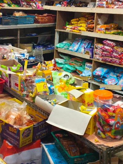 The image displays the interior of a convenience store located in Pinnwe, Myanmar. The scene is characterized by multiple shelving units and display surfaces densely stocked with various packaged consumer goods. On the left, metal shelves support blue and brown woven plastic baskets containing items, with additional packaged goods and a clear plastic bottle on lower shelves. The foreground and mid-ground feature a counter or table surface crowded with open cardboard boxes serving as product displays. These boxes contain individually wrapped candies, snack bags, and other small packaged foodstuffs, including a box for "Teh Gelas 250 ml" visible in the lower left. On the right, wooden shelving units are filled with rows of packaged snacks, instant noodles, and other processed food items in colorful plastic bags and boxes. A large clear plastic container with an orange lid, holding assorted candies, is positioned on the right-side counter. A green plastic basket containing dark-colored items is visible at the bottom right. No individuals are present in the depicted scene.