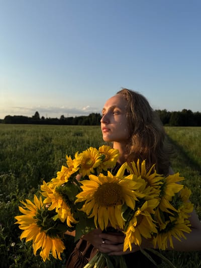 In a field at sunset in Aksaitovo, Russia, a young woman with blonde hair holds a large bouquet of sunflowers. She is wearing a dark dress and a ring on her left hand. The woman is looking off to the side with a thoughtful expression. The background consists of a grassy field and a line of trees in the distance, under a clear blue sky. The lighting suggests the golden hour.