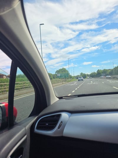 The image shows a view from inside a car traveling on a highway, likely the A66, in Thornaby-on-Tees, United Kingdom. The car is moving forward, with the dashboard and part of the passenger side visible in the foreground. The highway has multiple lanes, with other vehicles also in motion. Road signs indicate "Thornaby" and "A19 North & South Middlesbrough". The sky is blue with scattered white clouds, and there are streetlights visible along the side of the road. Trees and greenery line the highway. The overall scene depicts typical highway travel on a clear day.