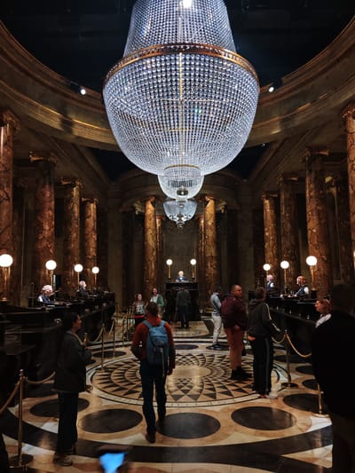 The image captures the interior of a grand hall, likely a set for a film or attraction, with a massive, multi-tiered crystal chandelier dominating the scene. The architecture features tall, marble-like columns and ornate gold-colored details around the upper walls and ceiling. The floor is patterned with concentric circles in black, white, and beige. Several people are present, standing in small groups or walking around. Some are standing behind podiums along the sides of the hall, possibly portraying characters. Ropes and stanchions are used to guide visitors. The location is possibly in Three Rivers, United Kingdom.