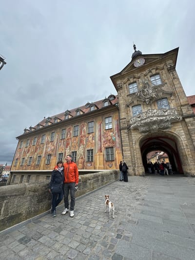 A slightly rotated medium-shot photograph captures two people standing closely together on the Old Bridge (Alte Brücke) in Bamberg, Germany, with the Altes Rathaus (Old Town Hall) as a notable backdrop.
The Altes Rathaus is a distinctive building with a half-timbered construction, adorned with paintings. It stands on an island in the middle of the Regnitz River. A clock is visible on the building's facade.
The people, a man and a woman, stand together on the bridge. The man is wearing an orange jacket and dark pants, while the woman is dressed in a black jacket and pants. A small dog stands near them on the cobblestone bridge. A group of people is passing through the archway of the Altes Rathaus.
The sky above appears overcast, suggesting cloudy weather.