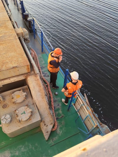 The image depicts two individuals on the deck of a vessel in the waters near Shestovo, Russia. Both individuals are wearing high-visibility orange vests and protective headwear. One person, wearing an orange helmet, is standing near the railing and appears to be speaking into a handheld device. The other person, wearing a white helmet, is seated on a small structure with blue metal supports and wooden planks, looking out at the water. The deck of the vessel is painted green and shows signs of wear. Several metal hatches are visible on the left side of the image, along with a thick, coiled rope. The water is dark and exhibits ripples, suggesting movement. The overall lighting indicates it is daytime, possibly late afternoon or early morning, given the warm tone of the sunlight on the vessel's structures.