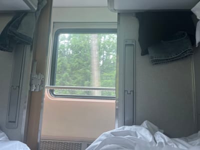 The image depicts the interior of a train compartment. A window reveals a blurred view of green trees, suggesting movement. Inside, a metal bar runs horizontally across the lower portion of the window. On the left, a pair of denim jeans hangs from a hook. On the right, another pair of folded denim jeans is visible above a black garment. A white duvet or comforter is prominent in the foreground, partially obscuring the lower section of the compartment. The walls of the compartment are textured grey. A metallic handle is present on the right wall. The scene appears to be a sleeping berth on a train. The image does not provide specific details about the city of Sloboda, Russia.