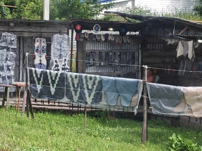 This image depicts an outdoor stall in Novye Vyselki, Russia, featuring various items for sale, including clothing and what appear to be household textiles. The stall is constructed with corrugated metal and wood, with a tarp-like roof providing shelter. On the left side of the stall, several pieces of outerwear, possibly vests or jackets, are displayed on hangers. They have a grey, patterned design. In the center, a colorful vest with floral patterns and traditional fasteners is hung, with additional items below it. To the right, more clothing items and some circular objects, possibly hats or decorative items with symbols, are visible. Shelves behind the stall display rows of what appear to be small containers or bottles. In the foreground, several large textiles with camouflage and blue patterned designs are draped over a railing, suggesting they are also for sale. The ground in front of the stall is covered in green grass with scattered small white and yellow wildflowers. A person, partially visible, is seated behind the stall, with their upper body in view. The overall scene suggests a small roadside market.