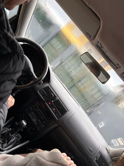 The image depicts the interior of a vehicle, likely a Volkswagen Golf Mk4 based on dashboard design, from a tilted perspective looking forward through the windshield. A person, partially visible on the left, wears a dark, possibly padded jacket, with their right hand on the steering wheel. The steering wheel rim appears worn. The dashboard is dark grey, featuring a center console with a radio/CD player, climate controls, and air vents. A red hazard light button is visible, and an amber indicator light, possibly for engine management or a low fluid warning, is illuminated on the dashboard. A mobile phone holder is clipped to an air vent. A rearview mirror is mounted centrally, reflecting a blurred interior or exterior. In the lower right of the frame, the bare feet of a second individual, a passenger, are visible, covered by a light-colored blanket or garment, with a ring on one toe.

Through the water-splattered windshield, the exterior environment is identifiable as an automated car wash bay. Green horizontal elements, consistent with rollers or brushes, are prominent. A bright yellow structure or light source is visible in the upper right background beyond these elements. Dark, wet surfaces are visible below the green elements. The scene suggests the vehicle is stationary within the car wash. This scene is located in Santa Maria da Feira, Portugal.