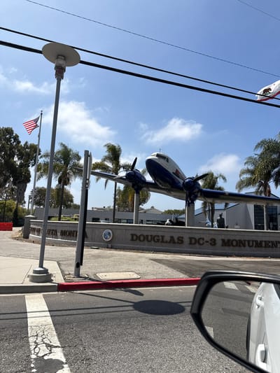 A monumental Douglas DC-3 airplane is on display at the Santa Monica Airport in Santa Monica, California, USA. The airplane is painted blue and white and is elevated on two pylons. A plaque in front of the airplane reads "DOUGLAS DC-3 MONUMENT". The airport building is visible in the background, along with palm trees and a clear blue sky with some clouds. An American flag and a California state flag are flying on poles to the left of the monument. The image is taken from the perspective of someone in a car, with the side mirror reflecting the road and some of the surrounding environment.