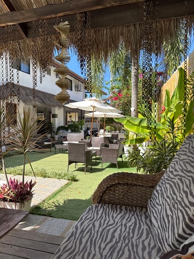 This image depicts an outdoor leisure area within a property in Panglao, Philippines. The scene is viewed from under a thatched roof structure, featuring decorative hanging metal chains and a spiral wooden wind chime.

In the immediate foreground, a dark brown woven rattan chair with a grey and white zebra-print cushion is visible on the lower right, with a wooden surface partially seen on the lower left.

The midground features a meticulously maintained green lawn. A light-colored stone path with embedded dark pebbles traverses the left side of the lawn. Several outdoor seating arrangements are placed on the grass, each consisting of multiple dark brown wicker chairs and matching low tables. Two large white market umbrellas provide shade over some of these furniture sets. Potted plants, including a dracaena with reddish-brown leaves, are arranged along the path.

In the background, a two-story white building with a thatched roof section and dark-framed windows stands on the left. An air conditioning unit is mounted on the building's exterior. To the right, dense tropical vegetation, including tall palm trees, broad banana leaves, and flowering bushes with red and pink blossoms, creates a lush backdrop. A vertical bamboo fence is visible on the far right behind the foliage.

The environment indicates a bright, clear day with strong sunlight, evidenced by distinct shadows. No individuals are present within the frame.