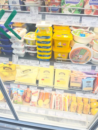 This media file shows a refrigerated display case filled with various types of cheese and dairy products in a supermarket. The products are predominantly packaged in yellow, white, and blue, with some featuring images of food or branding. Visible text on the labels is in Cyrillic, indicating Russian or Belarusian language. Brands like "Hochland," "LANDERS," and "Брест-Литовск" are present. Specific product descriptions include "Крем творожный" (cream cheese), "Сыр плавленый" (processed cheese), and various types of "Сыр" (cheese) with different flavors and fat percentages.

The display is well-stocked, suggesting a recent restocking or a busy shopping environment. The lighting is bright, typical of a retail setting. The overall scene depicts a typical grocery store aisle dedicated to dairy products. The time of day and weather are not discernible from the image. The location is identified as Navahrudak, Belarus, which aligns with the presence of Cyrillic text and brands common in that region. There are no people visible in the frame, focusing solely on the merchandise.