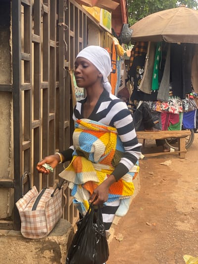 The image shows a woman in Barakin, Nigeria, standing in front of a stall filled with clothes. She is wearing a white headscarf and a black and white striped sweater, and is holding a colorful patterned blanket and a black plastic bag. Her right hand is holding a folded banknote, appearing to be a 500-naira note. Beside her, on a metal structure, is a plaid suitcase. Behind her, the stall displays various clothing items, including t-shirts, jeans, and dresses, under a makeshift awning. The ground is made of reddish-brown earth. The overall scene suggests an outdoor market setting. The lighting indicates it is daytime, likely under a bright sky. The woman's expression is focused, possibly on a transaction or organizing her goods. Visible text on the banknote reads "500".