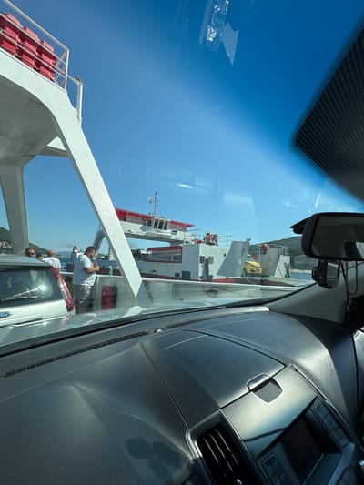 The image depicts a scene from inside a vehicle, looking out through the windshield and side window. The vehicle appears to be on a ferry, as indicated by the large ship visible in the background and the deck structure in the foreground. The sky is a clear, bright blue, suggesting a sunny day.
The main subjects are several large ships docked or in the water. One prominent ship is white with red accents and has multiple decks. Life rafts, stacked in bright red containers, are visible on the ferry's railing. In the middle ground, there are people on the ferry deck. One man, wearing a white t-shirt and light-colored shorts, stands near a vehicle and appears to be smoking. Other individuals are also visible near the vehicles. The vehicles themselves, including a white van with "Ford" visible on its rear, are positioned on the ferry.
The scene is illuminated by bright sunlight, casting strong shadows and creating highlights on the car's dashboard and the ferry's structure. The time of day appears to be midday, given the intensity of the sunlight and the clear sky. The setting is a coastal area, with buildings and hills visible in the distance behind the ships. The overall impression is one of a travel day, possibly a ferry crossing, with a bright and clear day for the journey. The image is oriented vertically, as if the camera was held in a portrait orientation.