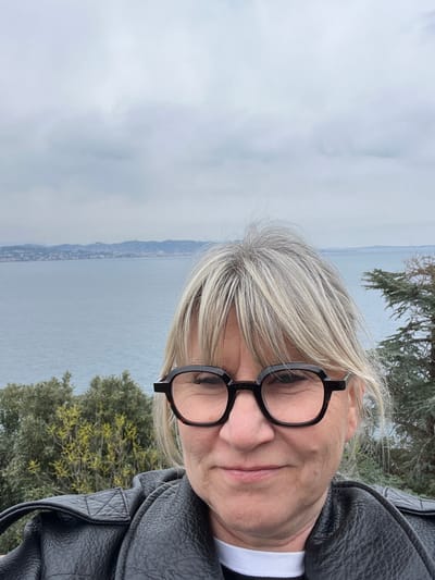 An adult female is depicted in a self-portrait photograph taken outdoors, overlooking a coastal scene in Théoule-sur-Mer, France. The subject, positioned centrally and filling the lower half of the frame, has light skin, medium-length blonde and grey hair with bangs, and wears dark, thick-rimmed eyeglasses. She is dressed in a black, textured jacket, possibly of faux leather, over a dark top with a visible white horizontal stripe at the collar. Her facial expression shows a slight, closed-lip smile with narrowed eyes, indicating a relaxed demeanor.

The background reveals an expansive body of blue-grey water, consistent with the Mediterranean Sea, stretching across the mid-ground. A distant coastline featuring low-lying hills and scattered structures is visible on the horizon. The sky above is overcast, presenting a uniform light grey cloud cover. In the immediate foreground, to the left and right of the subject, natural vegetation is present, including dense green shrubs and a darker green conifer-like tree, suggesting an elevated vantage point over the sea.