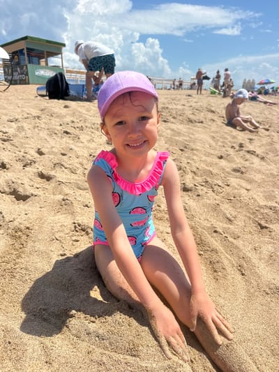 A young girl with blonde hair and a purple swim cap is lying on a sandy beach, smiling at the camera. She is wearing a blue swimsuit with a watermelon pattern and pink ruffles. Her arms are resting on the sand, which is scattered with footprints. In the background, other people are visible on the beach, some sitting or lying down, and others standing. There are umbrellas and beach chairs, suggesting a sunny day. A building with signage is partially visible on the left. The sky is blue with scattered white clouds. The scene appears to be a summer day at a beach resort. The building has text that appears to say "MUCHAKHO".