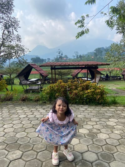 A young girl in a floral dress and pink shoes stands on a gray hexagonal-paved path. Her left hand holds the hem of her dress. In the background, there are open-air pavilions with red roofs and black benches on a green lawn. Lush vegetation, including trees and bushes, surrounds the pavilions. Mountains are visible in the distance under a partly cloudy sky. The location appears to be a park or recreational area, potentially in Penanggungan, Indonesia.