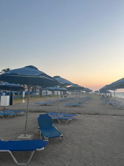 A wide-angle shot captures a beach in Rhodes, Greece, at dusk. Rows of blue lounge chairs and umbrellas are neatly arranged on a pebble beach. The sky transitions from a pale blue at the top to a soft orange and yellow hue near the horizon, indicating the setting sun. The sea is visible in the background, with a few people strolling along the shoreline. Palm trees and buildings can be seen behind the beach. The overall atmosphere is tranquil and serene.