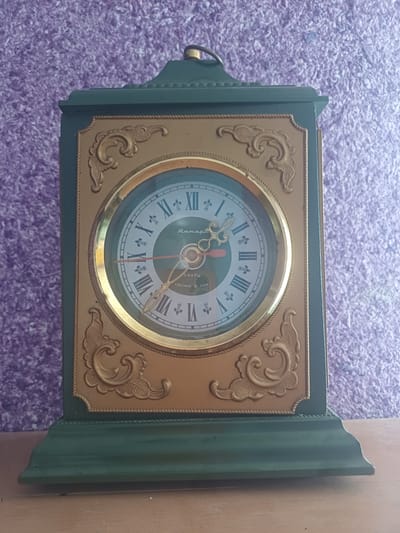 A dark green, rectangular mantle clock with a gold-colored face sits on a wooden surface against a textured purple background. The clock has Roman numerals and a brand name "Янтарь" (Yantar) written in cursive. The hands of the clock are red and gold. Below the brand name, it reads "КВАРЦ" (Quartz) and "СДЕЛАНО В СССР" (Made in USSR). The clock is decorated with ornate gold scrollwork on its face and a carved green base. The purple background appears to be a textured wall or fabric. The lighting suggests an indoor setting. There is no indication of the city of Jumashuy, Uzbekistan, in the image.