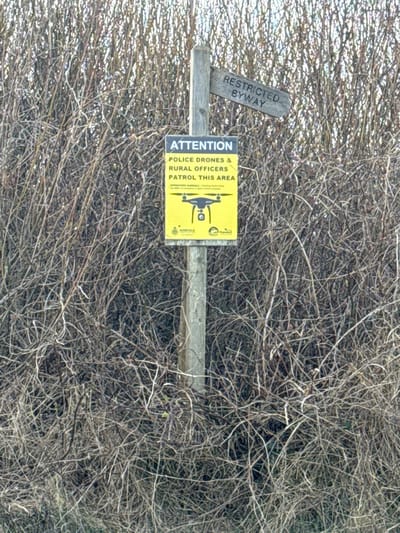 A weathered wooden signpost stands centrally, embedded in dense, tangled leafless vegetation typical of a rural hedge or scrubland. Two signs are affixed to the post. The upper sign is a small, rectangular piece of light-colored wood, angled upward to the right, bearing the faded inscription "RESTRICTED BYWAY" in black text. Below it, a larger, vertical rectangular sign in bright yellow displays prominent black text: "ATTENTION / POLICE DRONES & / RURAL OFFICERS / PATROL THIS AREA." Beneath this warning, an illustration of a black quadcopter drone with a camera is visible. Further small black text reads "OPERATION AMBERFIELD - Tackling Rural Crime" and "Help keep Norfolk a safe place to live and work." The bottom of the yellow sign features the "NORFOLK CONSTABULARY" logo on the left and a partially obscured logo (likely North Norfolk District Council) on the right. The vegetation background consists of numerous dark, intertwining bare branches and twigs, suggesting a winter or early spring season. The scene is located in a rural area within the North Norfolk district of the United Kingdom. No individuals or active interactions are depicted; the scene conveys informational warnings.