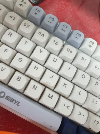 The image shows a close-up of a white and gray mechanical keyboard. The keyboard is angled, with some keys in focus and others slightly blurred. Visible keys include function keys (F4, F5, F6, F7, F8, F9), number keys (4, 5, 6, 7, 8, 9), and letter keys (R, T, Y, U, I, O, P, F, G, H, J, K, L, V, B, N, M). There are also symbols on some keys, such as dollar sign, ampersand, asterisk, parentheses, comma, period, semicolon, and question mark. The "SIBYL" logo is visible on the left side of the keyboard. The background appears to be a red, textured surface, possibly a desk or table. The keyboard is worn, with some dust or dirt visible between the keys. There is no indication of the location in Proyonangg, Indonesia.