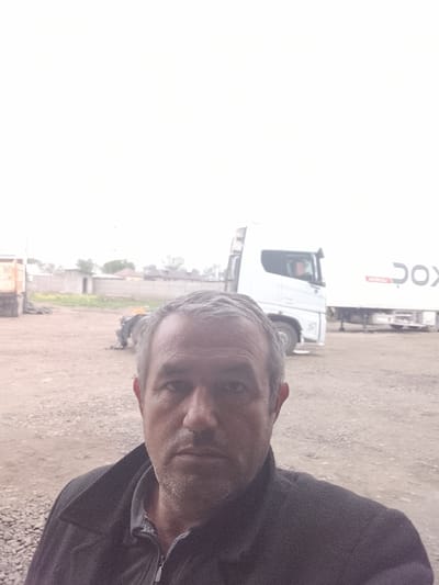 The image shows a middle-aged man taking a selfie. He has short, gray hair and a somewhat weathered face. He's wearing a dark jacket. Behind him is a gravel-covered area, with what appears to be a parked semi-truck with a white trailer. Further in the background, there is a partially obscured figure appearing to be working near some construction or storage materials. Also in the background are buildings with a compound-like structure. Based on the location provided, this scene is taking place in Sabir Rakhimov, Uzbekistan.